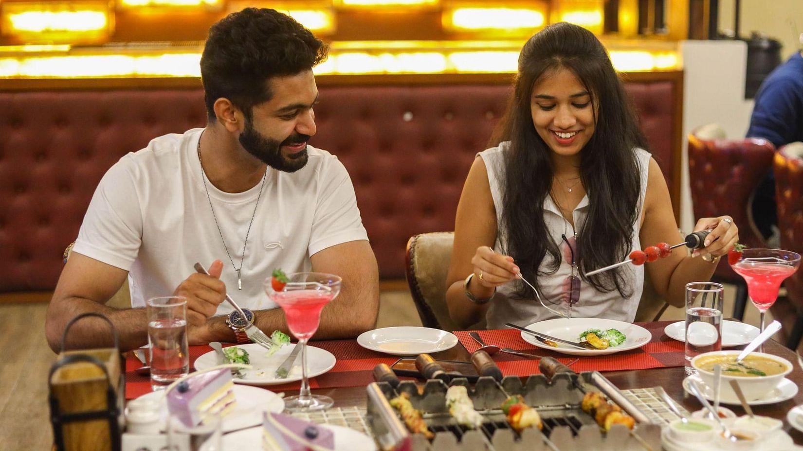 A man and a woman sit at a restaurant table, smiling and preparing to eat. Food and drinks are on the table, with skewers grilling in the center at Jenneys Residency