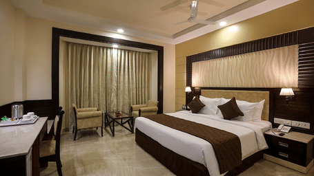 The room at Hotel Aaditya featuring a large bed, bedspreads, pillows, large windows covered with curtains, with a table, and a chair.