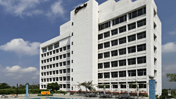 A large, modern white hotel building is shown behind a spacious blue swimming pool with lounge chairs on the patio - Clarks Avadh, Lucknow