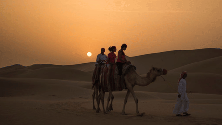 A small group of tourists riding a camel led by a local guide in the desert at sunset in Dubai, UAE.