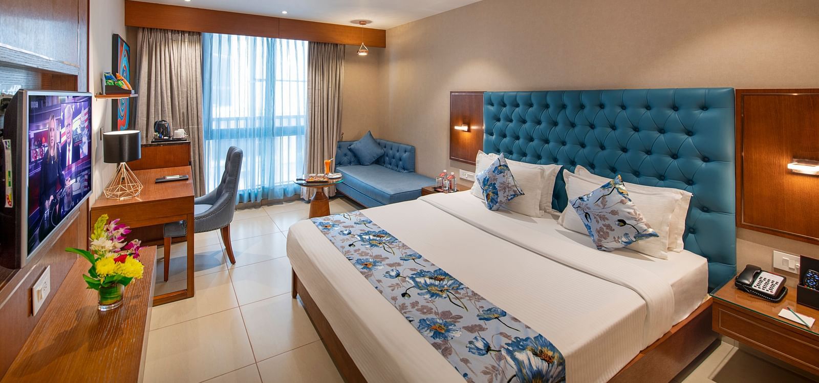 A well-appointed hotel room with a double bed with a blue headboard, a work desk, and a comfortable seating area by the window -  SK Lords Inn Ahmedabad
