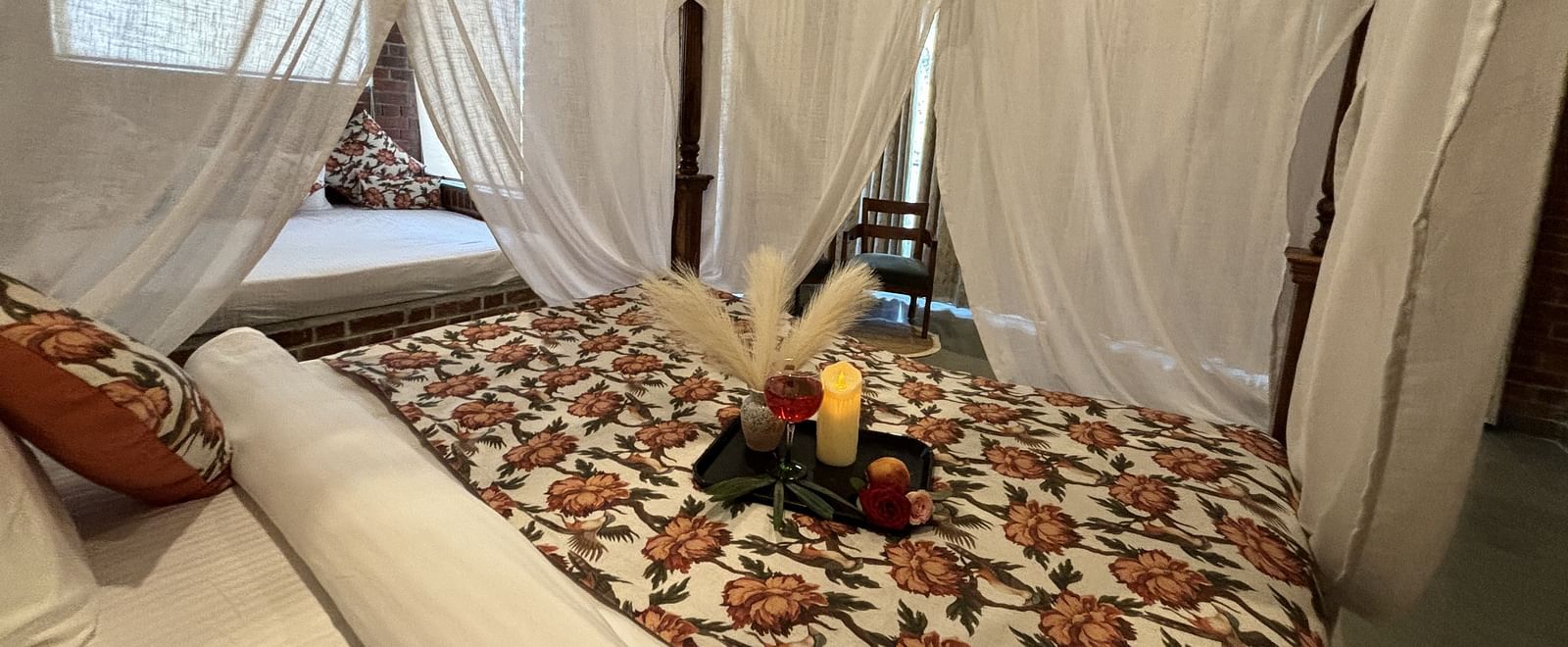 A bedroom at Tisya Farms, Gurgaon with a canopy bed, white curtains, and floral bedding with a tray of refreshments.