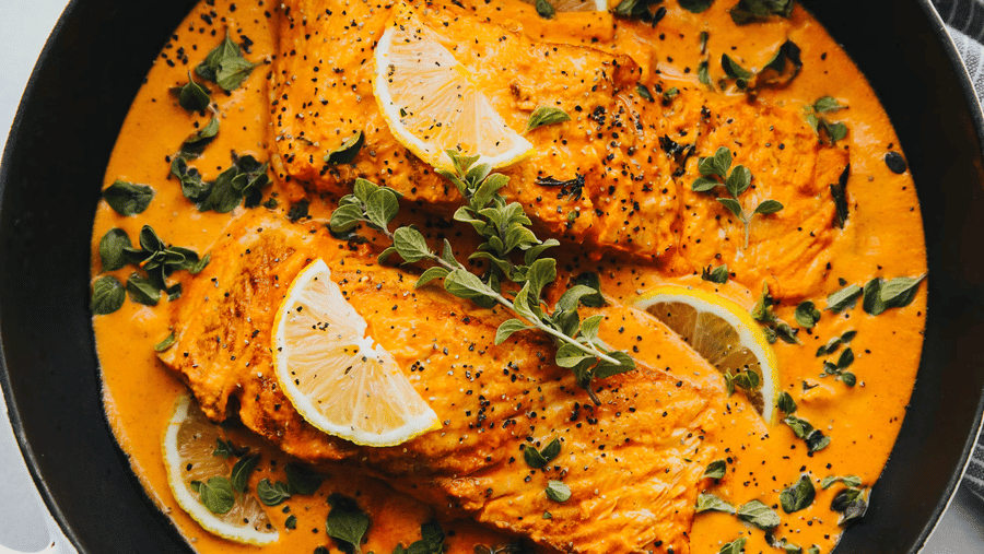Fish fillets in a creamy orange curry sauce garnished with lemon wedges, cracked black pepper, and fresh herbs, served in a black pan.