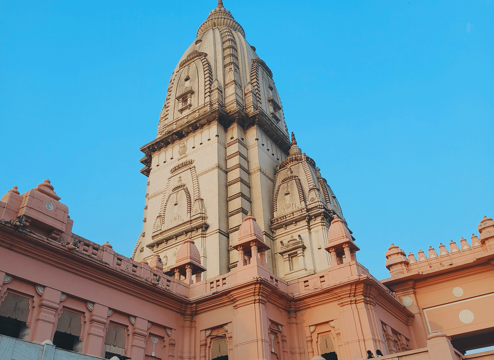 Tall cream stone spire of the New Vishwanath Temple at BHU campus in Varanasi rising above pink sandstone walls and a pillared courtyard under a clear blue sky.
