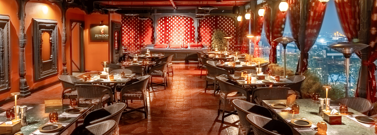 An indoor restaurant with tables, cushioned seating, textured ceiling, and large windows with a cityscape view at night at Infiniti Hotel, Indore.