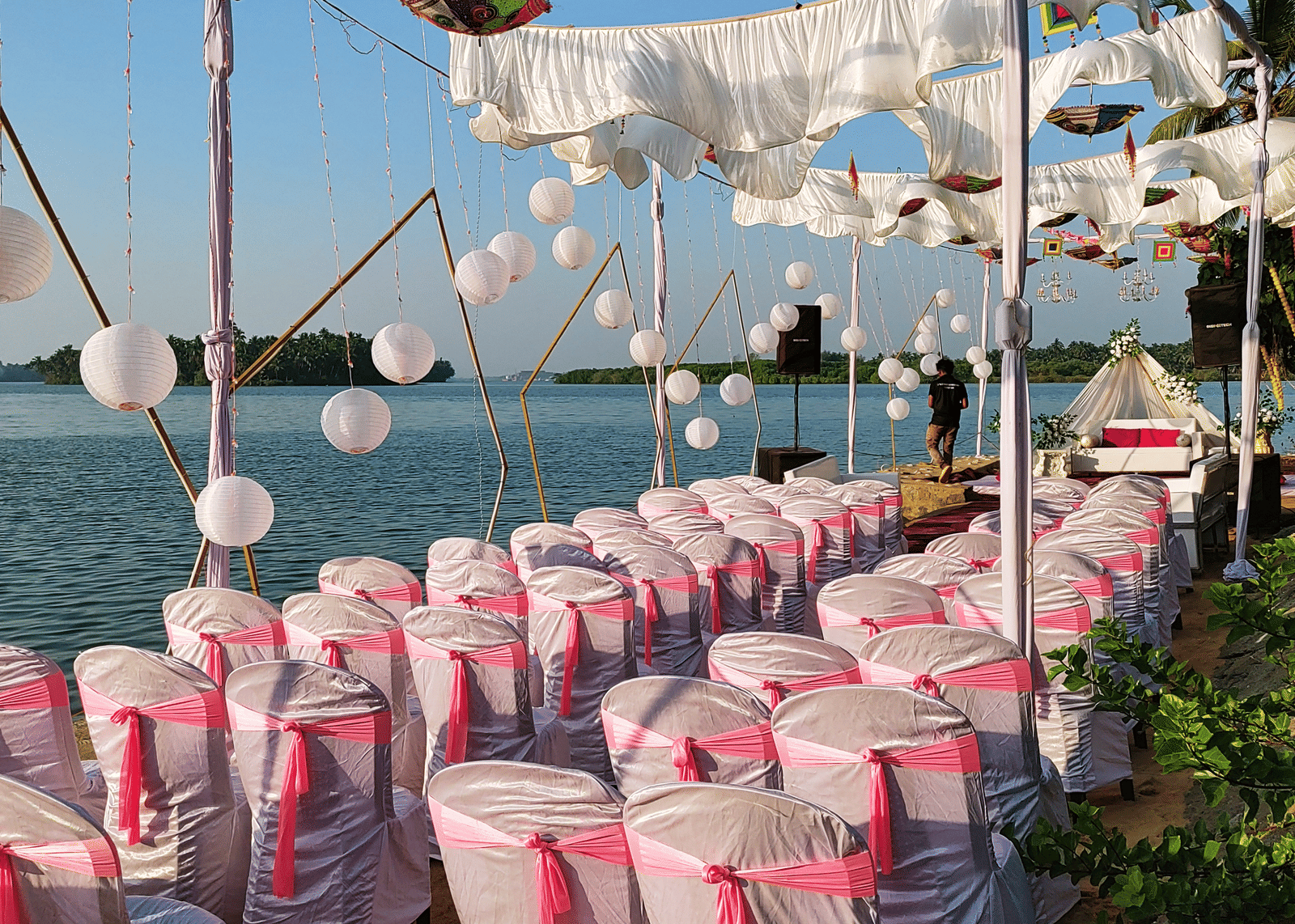 Rows of white chairs with pink covers arranged outdoors under hanging umbrellas at Paradise Lagoon Resort, Udupi.