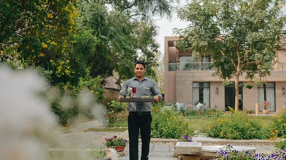 Man in black uniform serving drinks in an outdoor garden with buildings in the background at Karma Chalets.