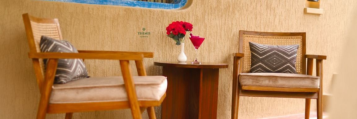 a balcony of classic villa decked with two chairs and a side table at a view of classic Villas with a plush bed and wooden furnishing at interior of Classic Villas arrayed with a sofa, trinkets and a lamp hanging from the ceiling at Themis Mudhouse - A Nat