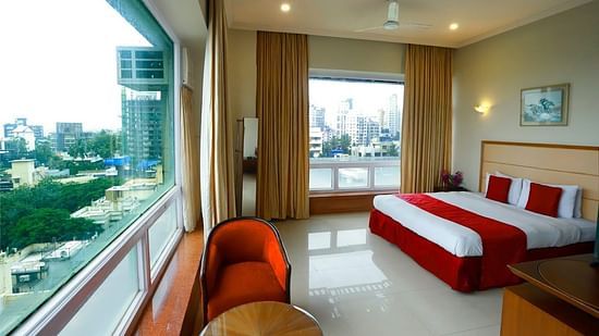 A room adorned with a well-made bed, a coffee table, a red sofa chair, and two large windows offering an expansive city view.