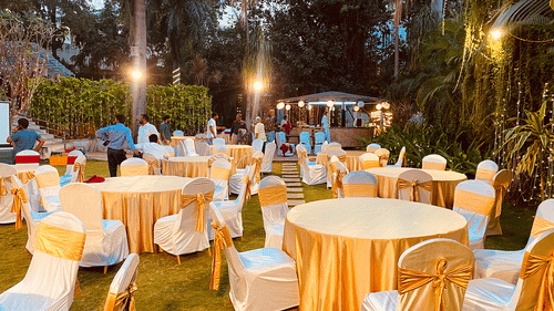 Outdoor event setup with round tables covered in white and gold cloths, surrounded by chairs under string lights.