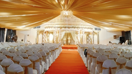 Lavishly decorated indoor venue with a red carpet aisle, white-covered chairs, and a golden canopy.