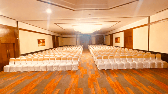 Indoor hall with rows of chairs and a stage area, featuring a wooden floor and warm lighting.
