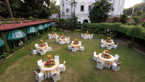 Round table seating arrangement on the lawn area at Kenilworth Hotel, Kolkata, with trees and buildings surrounding the lawn area. 