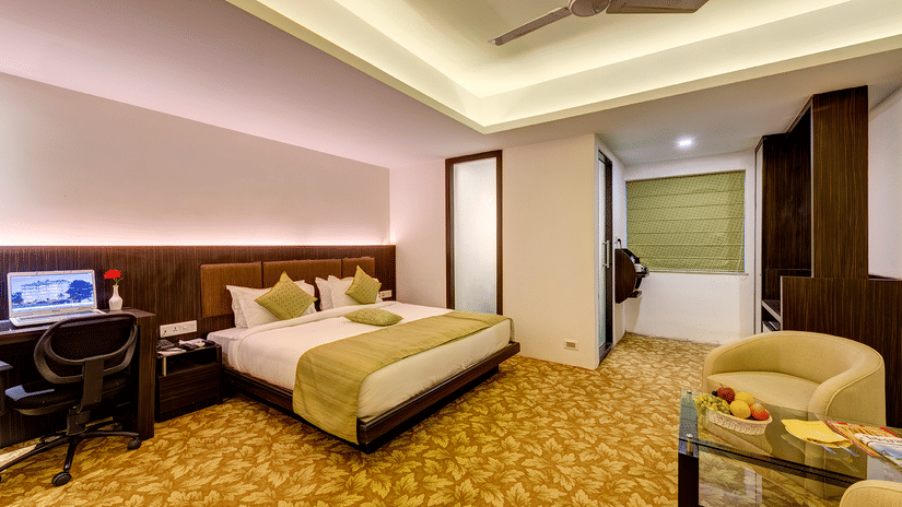 Premium room with king bed, green accents, work desk with monitor, red flower vase, armchairs with fruit table, patterned carpet, and warm lighting at Viceroy Boutique Hotel, Kolkata.