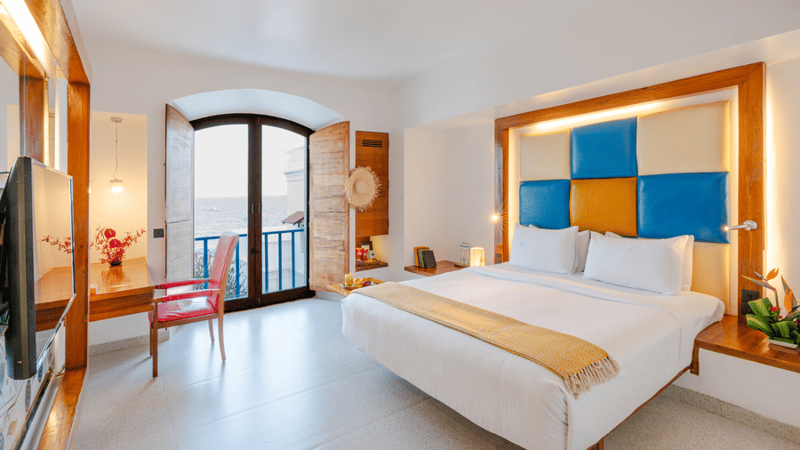 Wide view of a room at The Promenade Pondicherry with a king bed, blue and yellow headboard, wall-mounted TV, balcony access, and bright natural light.