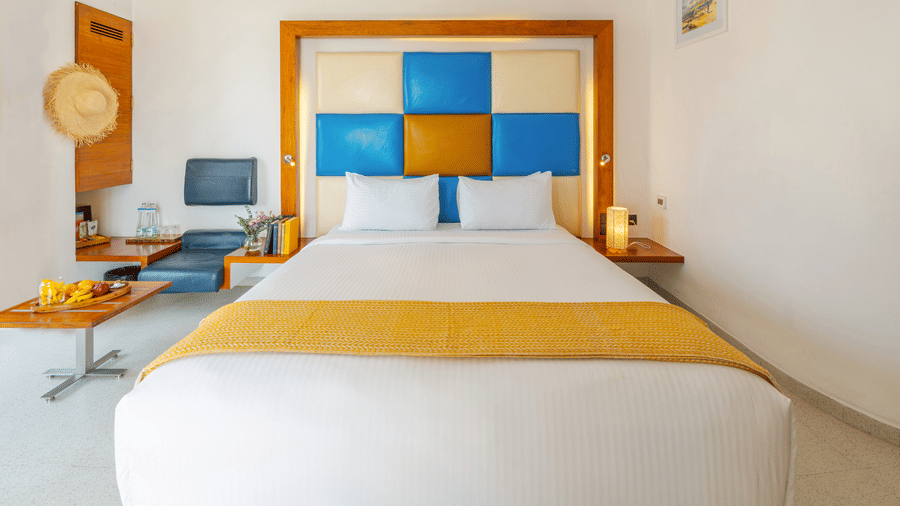 Side view of a room at The Promenade Pondicherry with a king bed, blue and yellow headboard panel, bedside lamps, work desk, and soft ambient lighting.