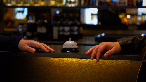 Two pairs of hands rest on a dimly lit bar counter with a silver service bell in the centre, bottles blurred in the background.