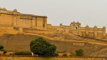 A grand, ancient amber-coloured fort perched on a hill, overlooking a body of water with lush greenery