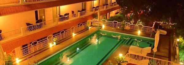 A swimming pool with a lounge at poolside and surrounded by room balconies and lights in the evening at Hotel Mamallaa Heritage, Mahabalipuram.