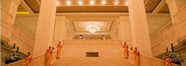 A wide staircase with railings, ceiling lights and hotel staff on either side of the stairs in the lobby area at Hotel Mamallaa Heritage, Mahabalipuram.
