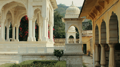 A white domed structure stands next to a walkway with arched colonnades, under a clear sky.