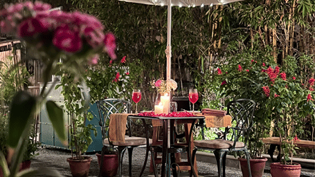 Under the umbrella nook at Karma Chalets featuring a cute dining setup for 2 under an umbrella with string lights placed amidst flower vases and tall trees on a small chequered floor.
