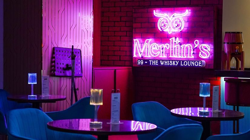 blue chairs and wooden table placed on a marbled floor with merlin's 99 signboard glowing in pink in the background