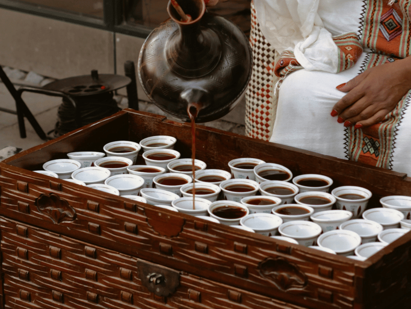 A person pouring coffee to numerous cups, kept inside a small table like structure in the outdoors