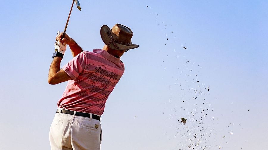 A man playing golf, captured mid-swing on a golf course, with green grass underfoot and a clear blue sky in the background.