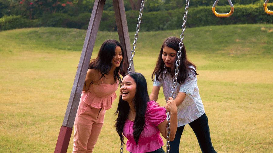 Three people playing at an outdoor swing set on a grassy field with trees in the background, one person seated on the swing and two standing.