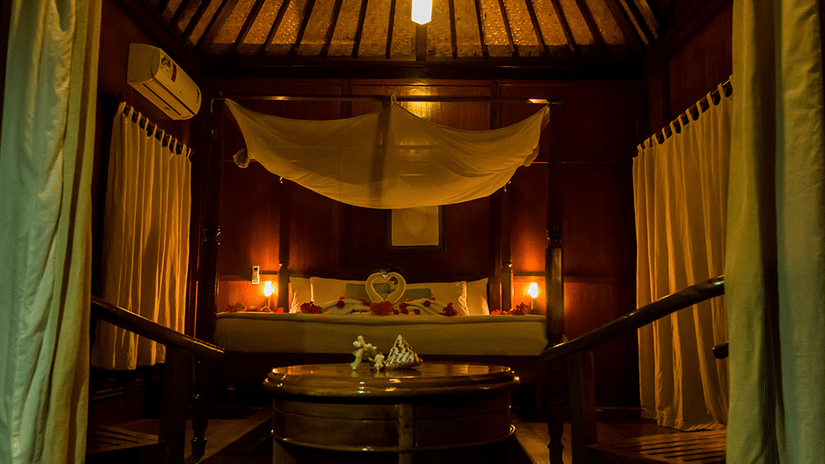 A close up view of the interior of Andaman Villas in Havelock Island with chairs, coffee table, a bed, AC, dove towels on the bed in view.