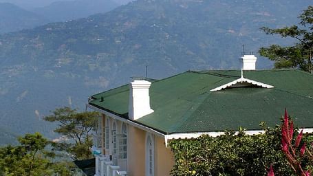 A white cottage with a green roof surrounded by flowers, set against a mountainous landscape with clear skies.