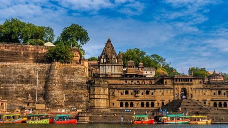 Historic riverside fort with boats docked along the water in Maheshwar, India, under a bright blue sky.