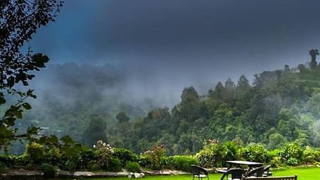 Lush green lawn and trees under a dramatic cloudy sky, with mountains in the background and a wooden bench in the foreground.