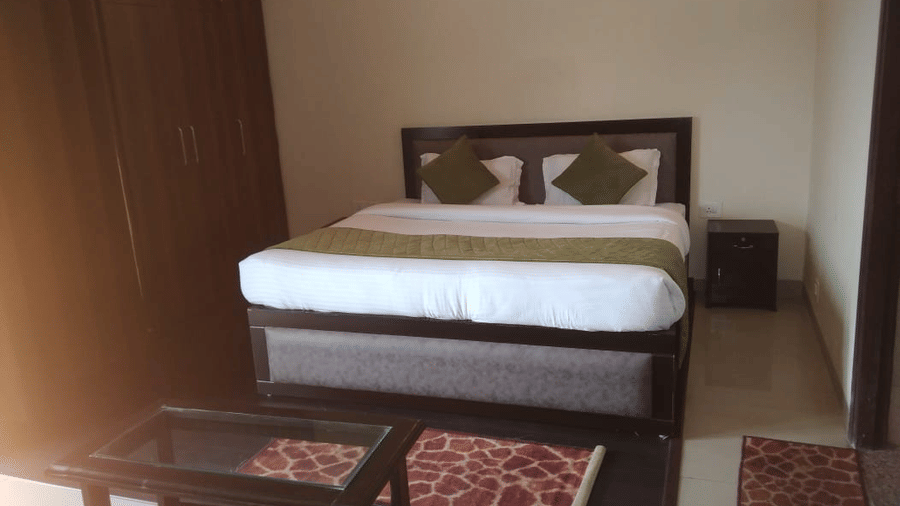 A dimly lit bedroom with a wooden double bed, a glass-topped table, red rug, and bedside table at Livbox, Rudrapur.