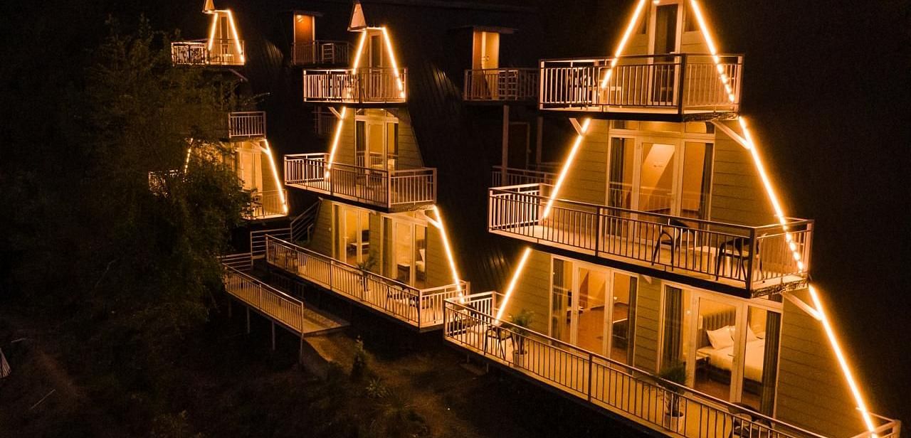 An aerial night view of a resort with several A-frame or pyramid-shaped cottages illuminated with warm lights.