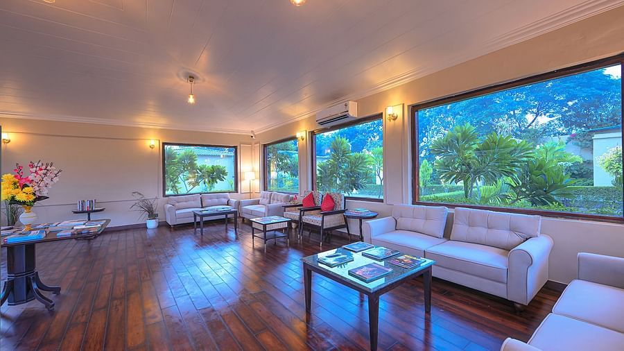 A well-lit lounge featuring plush white sofas, rich wooden flooring, and large windows overlooking lush greenery. Soft lighting and elegant decor create a warm and inviting atmosphere - Singhvilas, Ranthambore