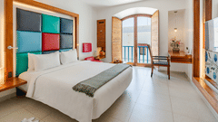 Wide view of a room at The Promenade Pondicherry with a king bed, multicoloured headboard panel, arched windows, balcony access, and natural light.