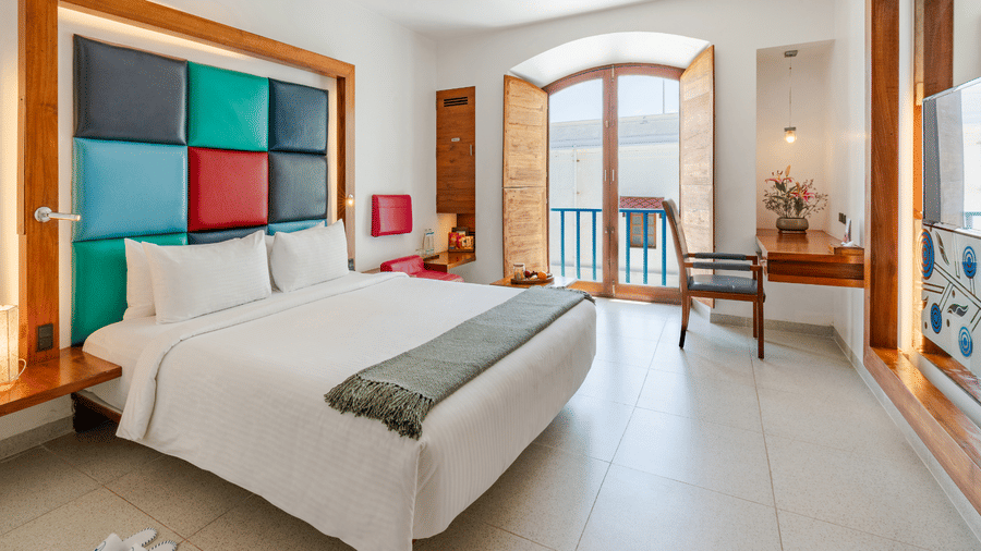 Wide view of a room at The Promenade Pondicherry with a king bed, multicoloured headboard panel, arched windows, balcony access, and natural light.