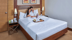 A couple sitting on a bed in a hotel room decorated for a honeymoon package, featuring food on the bed