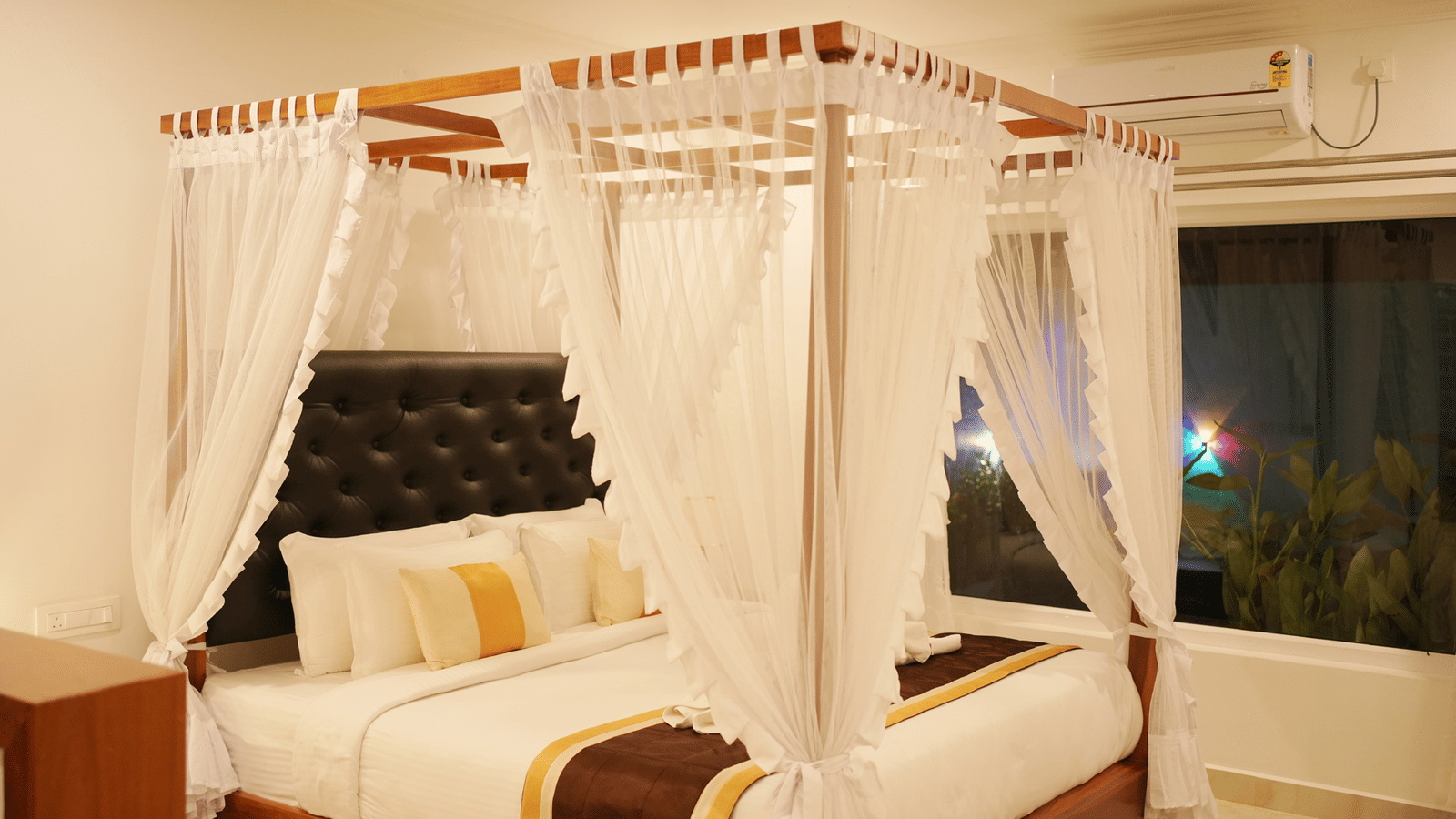 A modern bedroom featuring a four-poster canopy bed draped with white fabric and luxurious gold bedding at Coorg Orange Blossom Resort and Spa.