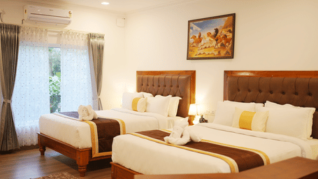 A spacious, well-lit hotel room featuring two large beds with upholstered headboards, brown runners, and towels folded like swans at Coorg Orange Blossom Resort and Spa.