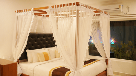 A modern bedroom featuring a four-poster canopy bed draped with white fabric and luxurious gold bedding at Coorg Orange Blossom Resort and Spa.