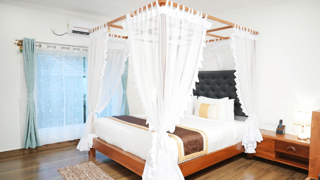 A luxurious hotel bedroom featuring a four-poster wooden bed with white drapery and a brightly lit window at Coorg Orange Blossom Resort & Spa.