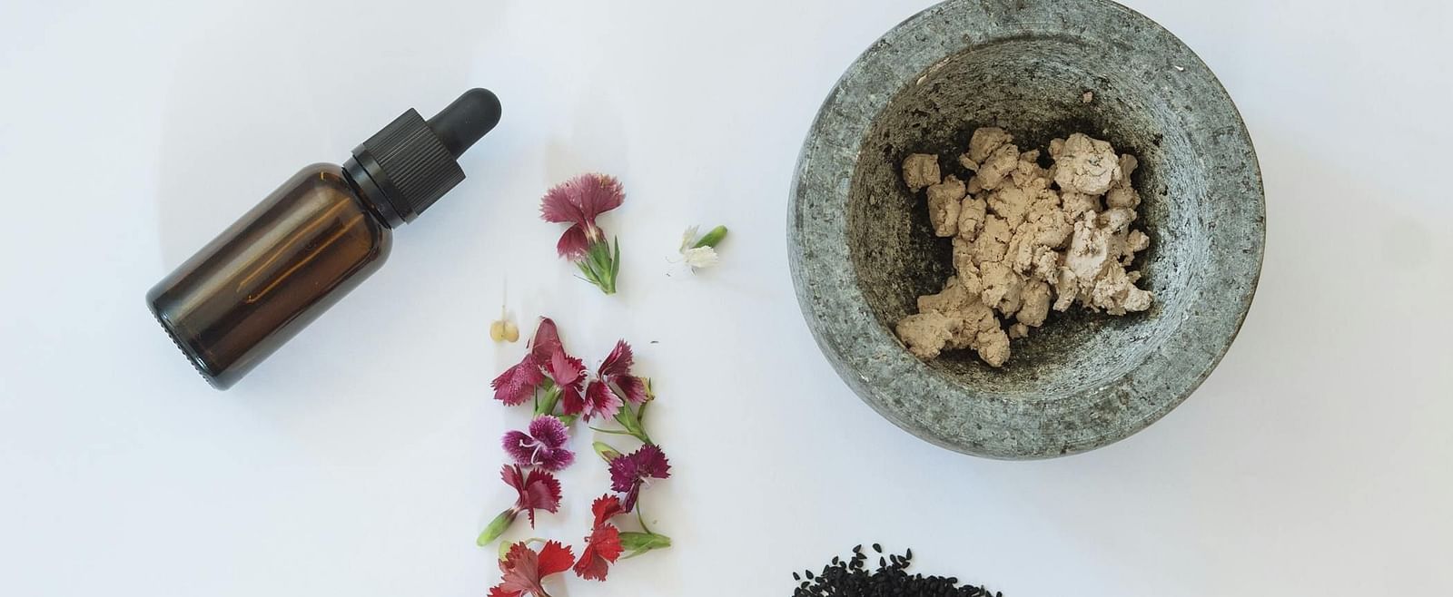 A glass vial, flowers, herbs and a mortar aesthetically placed on a white surface.
