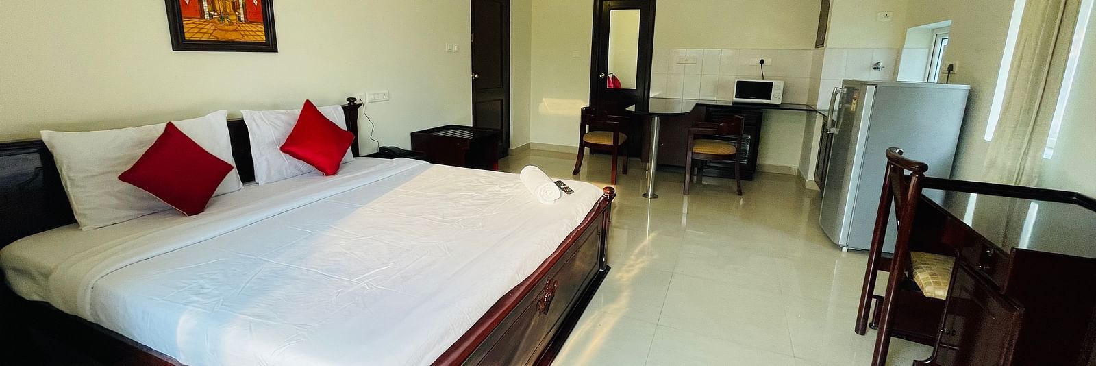 A room with a double bed featuring white linens and red cushions, a separate seating area, and a glimpse into a kitchenette space at Sanctum Suites Indiranagar, Bangalore.