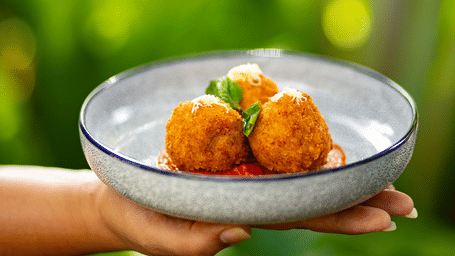 A person holding a small white bowl containing three golden-brown fried snacks garnished with herbs at Stanley Revelation.