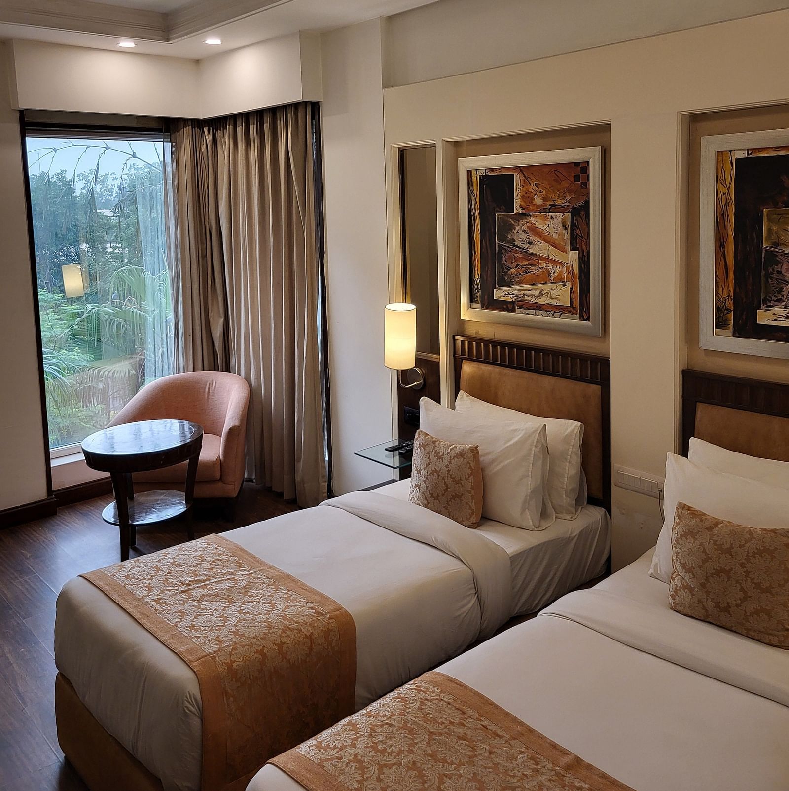 An image of a bedroom featuring twin bed, a sofa chair, a small centre table, bedside table with lamp, photo frames hanged on the wall, and a window o=overlooking the outdoors - Inde Hotel Signature Tower Gurgaon