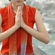 A woman in a red top sitting in a cross-legged position with her hands in prayer pose outdoors near a rocky river.