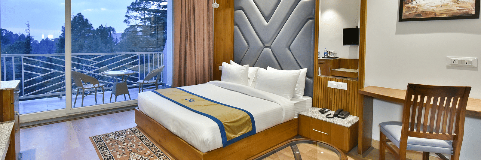Image of the luxury room with balcony with a bed, bed side tables, chair, work desk, and a glass door leading to the balcony with a scenic view in the background - DLS Ark Holiday Inn, Dalhousie
